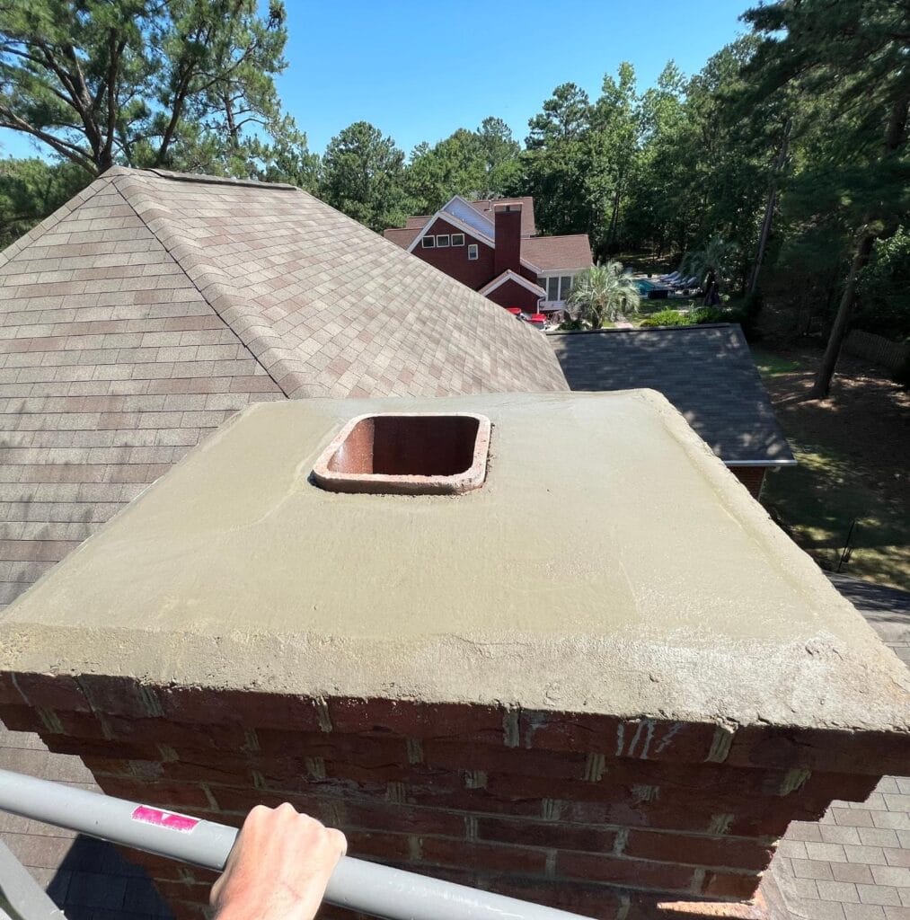 A view from a rooftop showing a brick chimney with a new concrete crown and a square flue opening, freshly serviced with professional Chimney Cleaning, surrounded by trees and nearby houses.