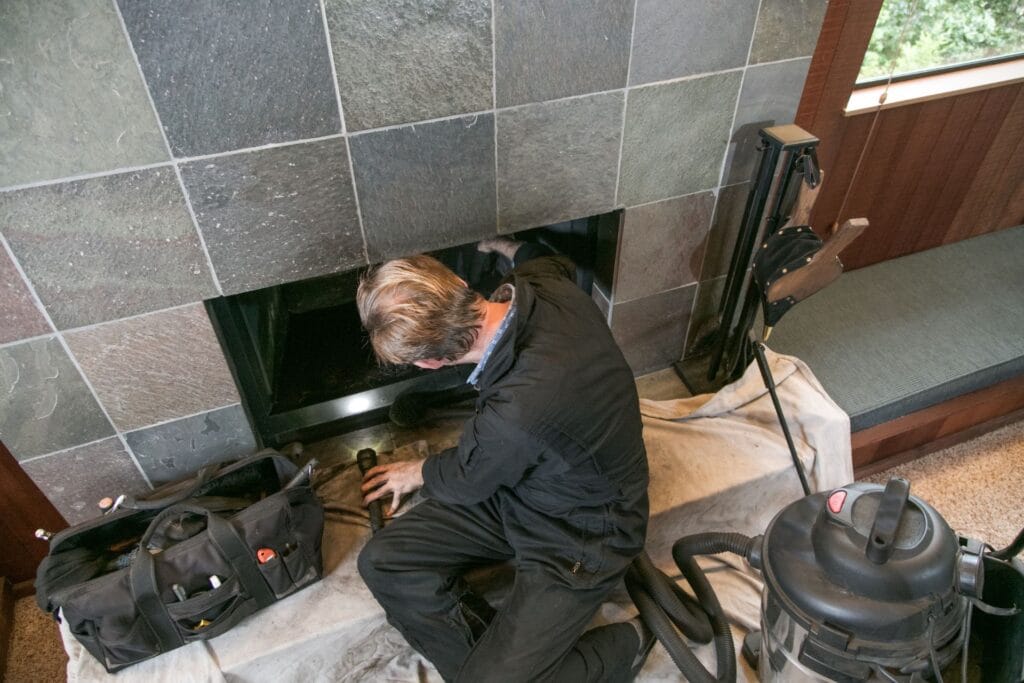 A person in work clothes cleans or inspects a fireplace as part of chimney repair, surrounded by tools and a vacuum, with a protective cloth spread on the floor.
