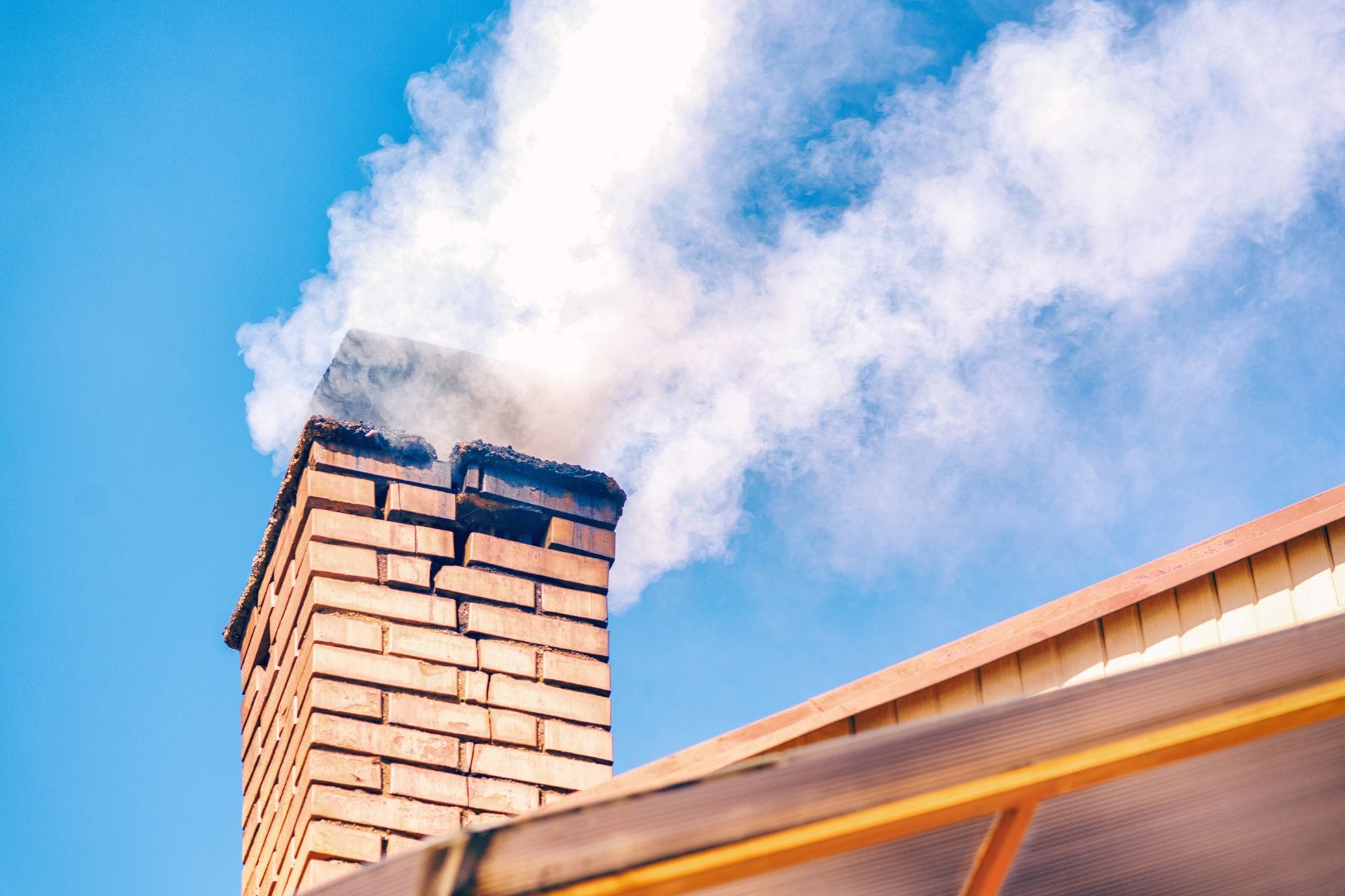 A brick chimney on a house roof emits white smoke against a clear blue sky, suggesting a recent visit from the chimney sweep.