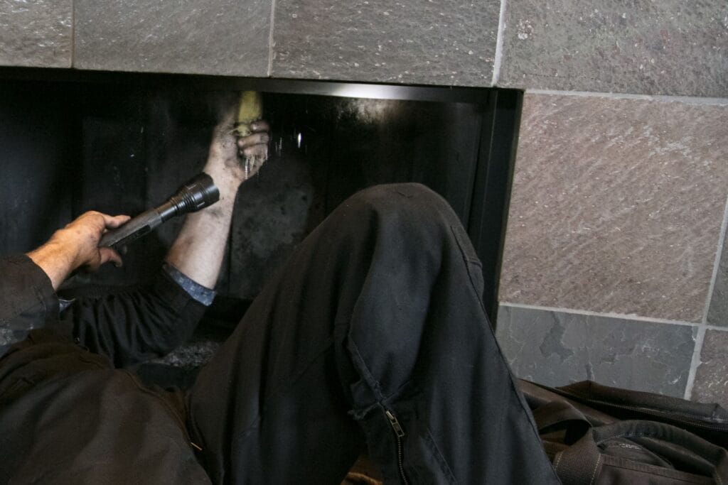 A person lies on the floor, performing a chimney inspection by cleaning the inside of a fireplace with a flashlight in one hand and their other hand inside the chimney.
