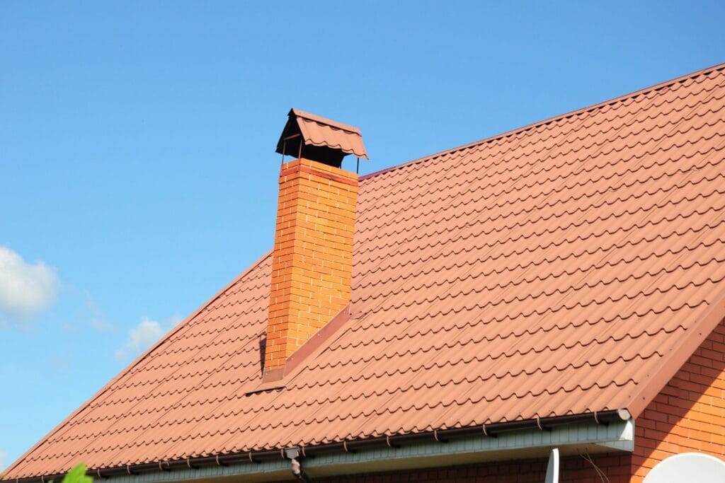 A brick chimney with a metal cap extends from a red tiled roof on a house under a clear blue sky, ready for expert Chimney Services.