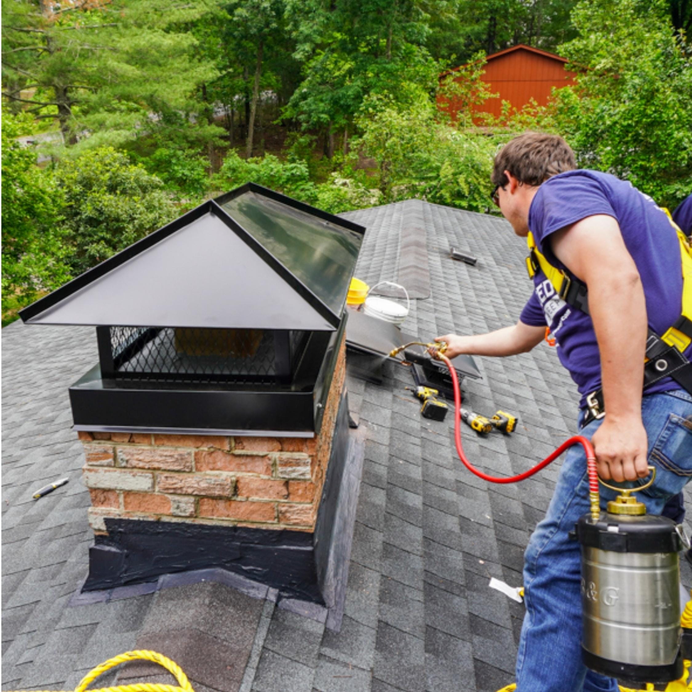 A worker wearing a safety harness inspects and performs maintenance on a brick chimney on a shingled roof, surrounded by trees.