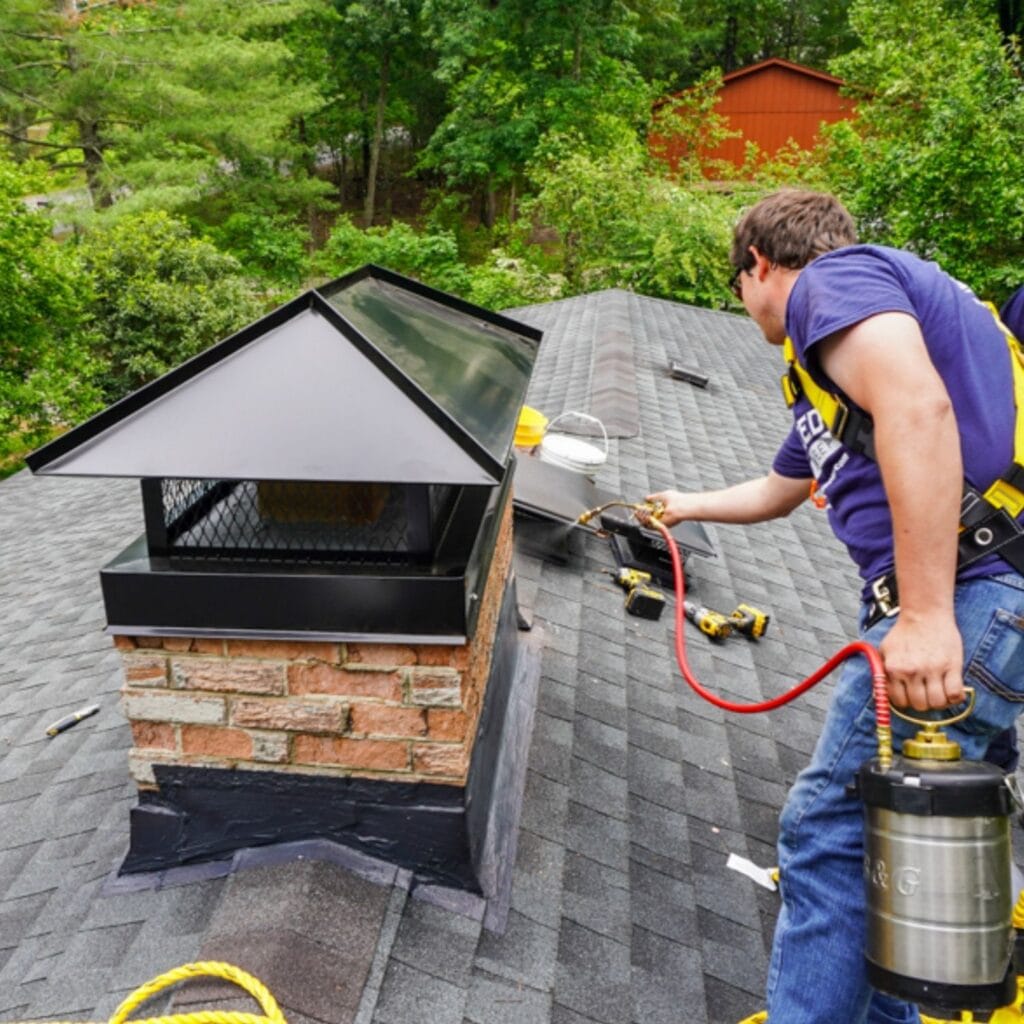 A worker in safety gear applies spray to a brick chimney cap on a shingled roof, with tools and greenery visible in the background.