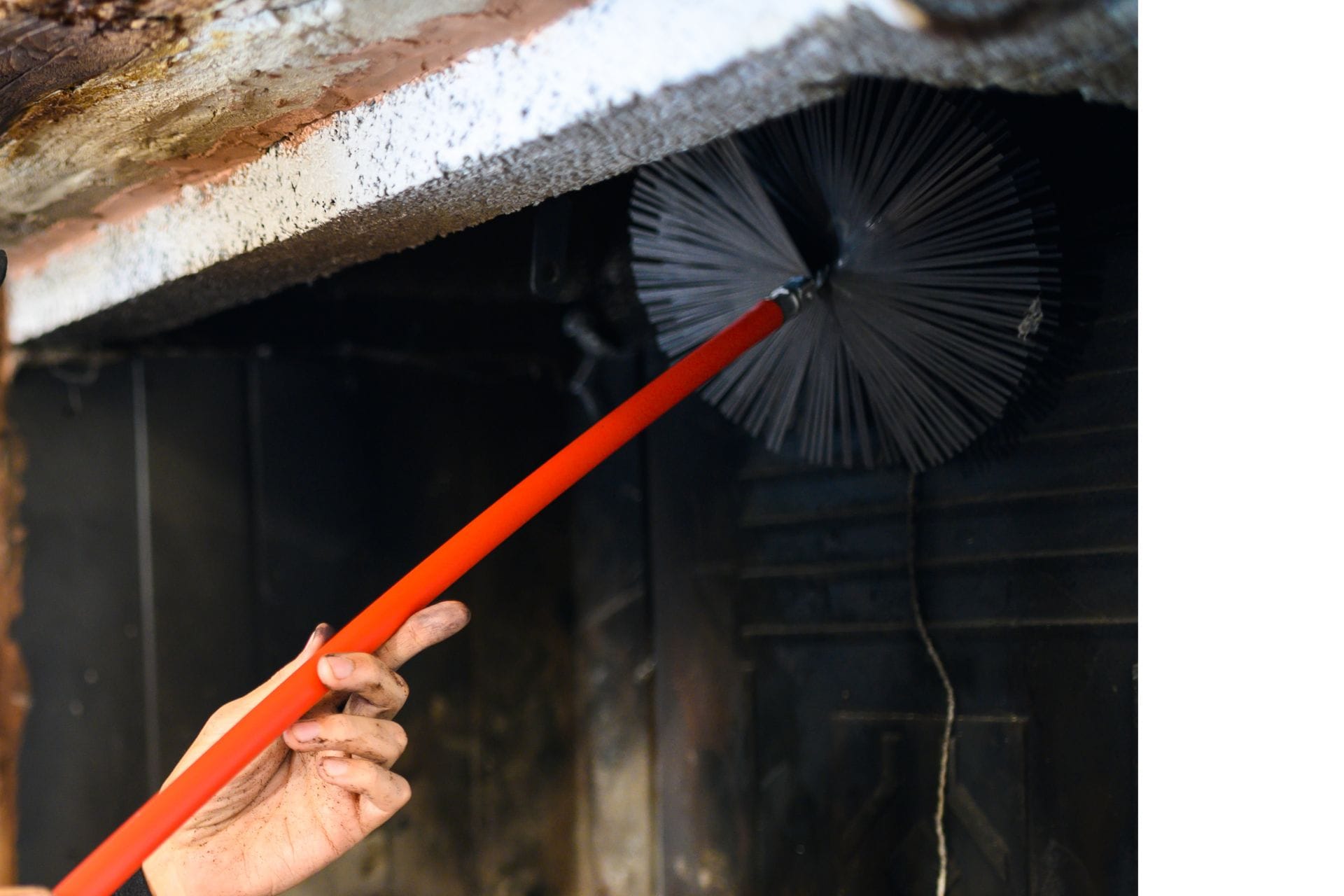 A chimney sweep uses a brush with a red handle to clean the inside of a fireplace, soot visible on their hand and the surrounding area.