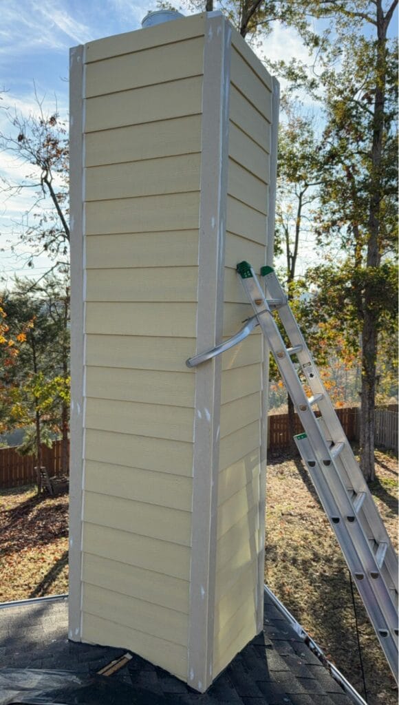 A tall, rectangular chimney covered in beige siding stands on a roof next to an extension ladder, with trees and a wooden fence in the background.