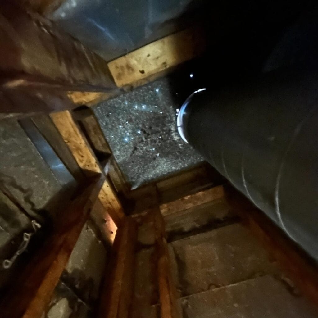 A view looking up a narrow shaft with wooden beams and a large black pipe, leading to an opening covered with debris and speckled light&mdash;ideal for illustrating the need for regular chimney cleaning or maintenance.
