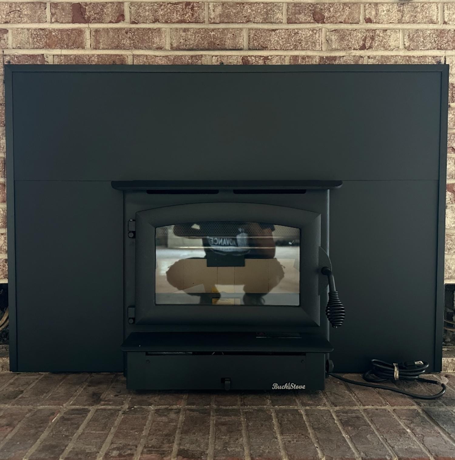 A black Buck Stove fireplace insert is installed against a brick wall, with visible power cords on the right side and a glass door on the front.