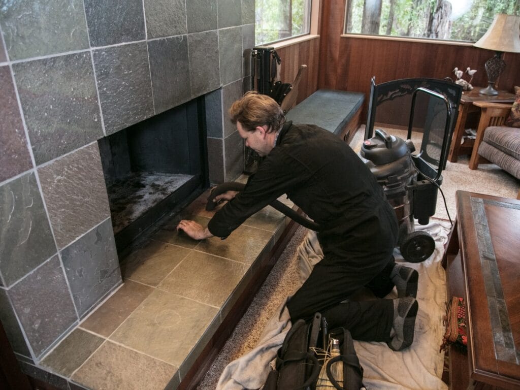 A person in work overalls kneels by a tiled fireplace, cleaning inside it with a vacuum cleaner and tools laid out nearby&mdash;highlighting the importance of regular chimney cleaning frequency for optimal safety and efficiency.