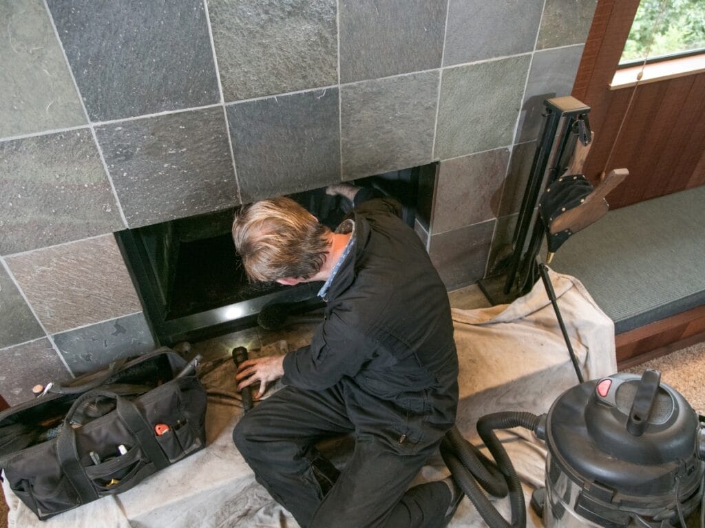 A person in work overalls cleans or inspects the inside of a fireplace, following a chimney inspection checklist, with tools and a vacuum nearby on a protective tarp.
