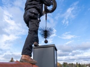 A person standing on a roof prepares to clean a chimney using a large brush attached to a flexible rod, following steps from a chimney inspection checklist.
