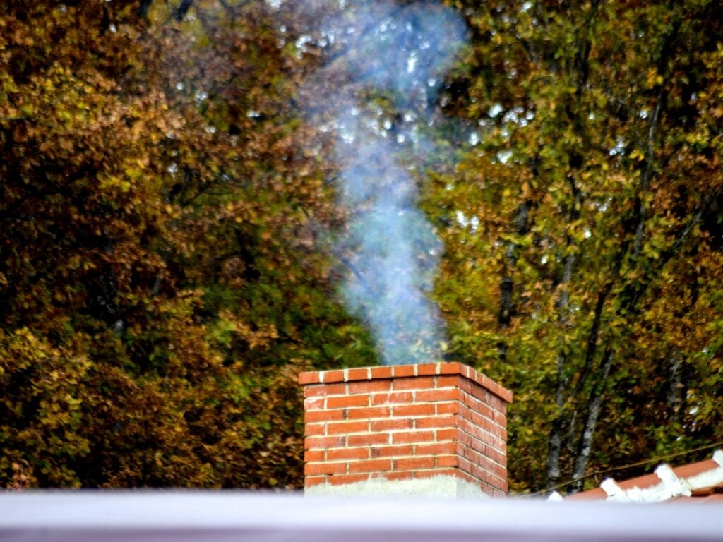 A brick chimney with quality chimney liners emits a stream of smoke, set against a backdrop of trees adorned with autumn-colored leaves.