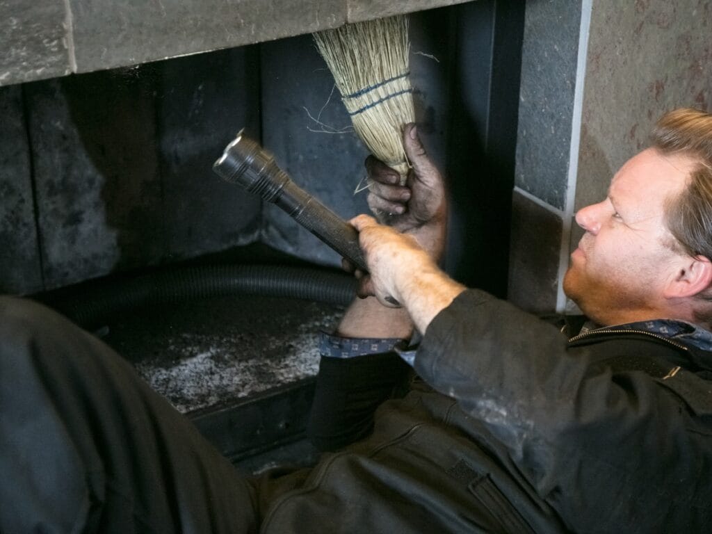 A person in work clothes lies on the ground, cleaning a fireplace chimney with a brush while holding a flashlight, inspecting the chimney liners for safety and cleanliness.