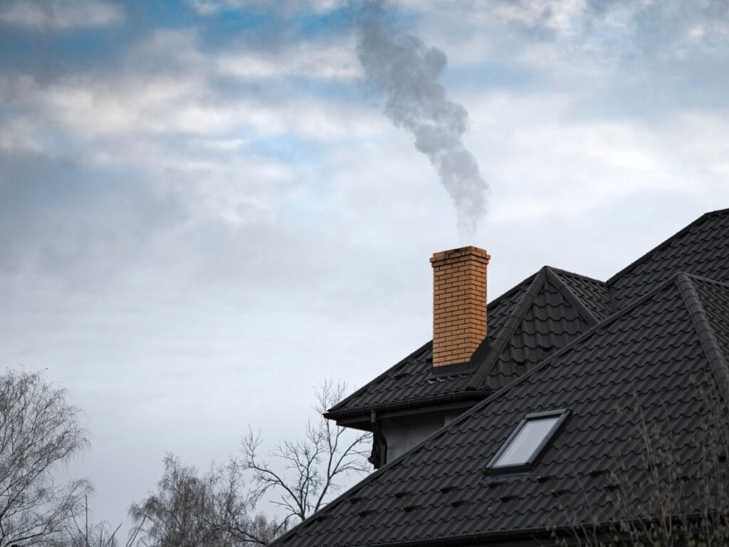 A brick chimney with sturdy chimney liners on a house roof emits smoke into a cloudy sky, with leafless trees visible in the background.