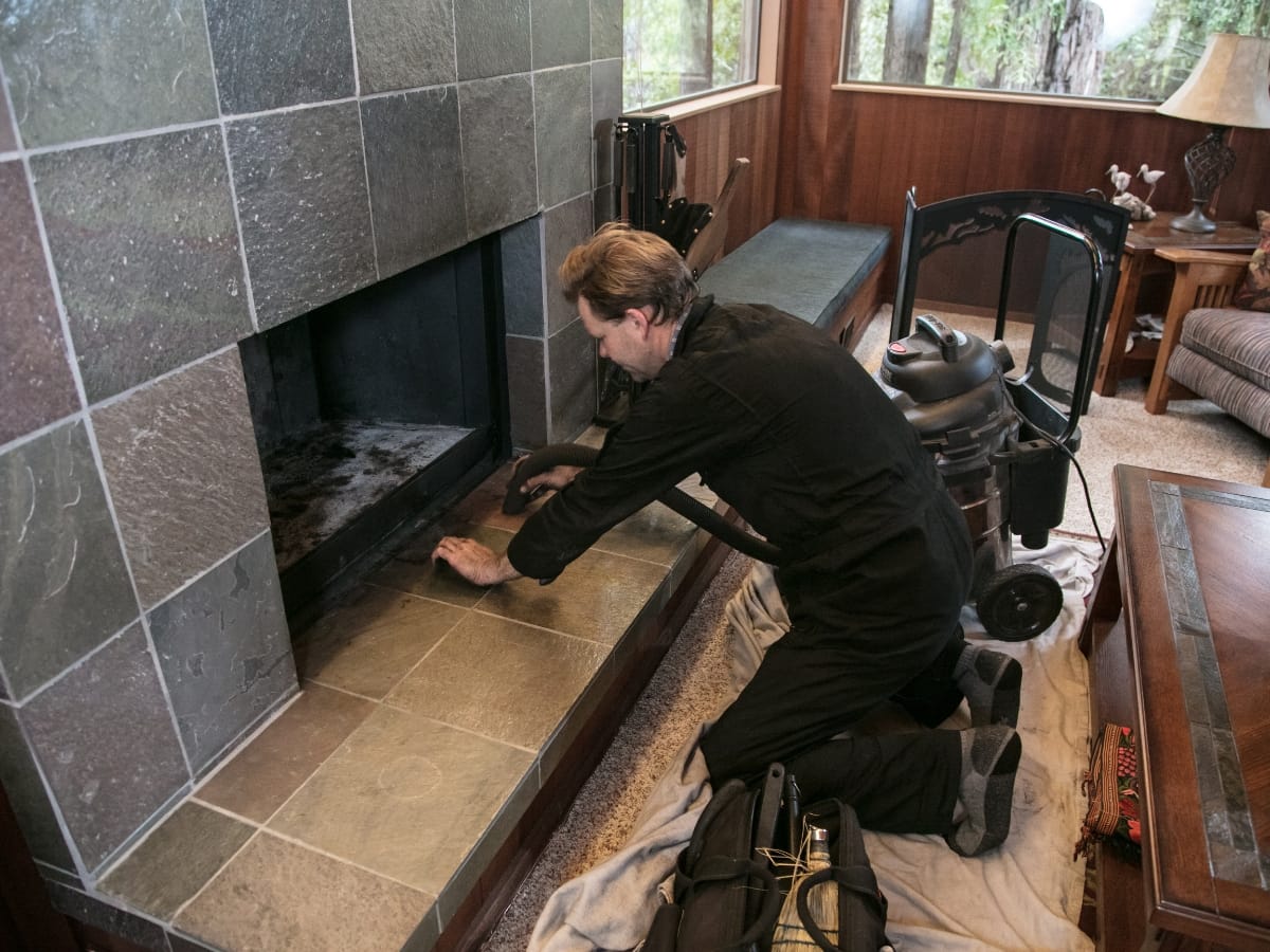 A person in dark clothing kneels on a protective cloth while cleaning a fireplace with a vacuum, ensuring the chimney liners remain free of debris in the living room.