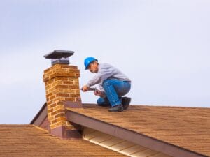 A person wearing a blue hard hat demonstrates tuckpointing explained, repairing a brick chimney while kneeling on a brown shingled roof under a cloudy sky.