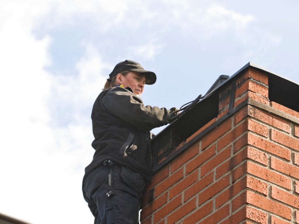Person in work uniform inspects or cleans a brick chimney on a rooftop under a partly cloudy sky, demonstrating key steps in Tuckpointing Explained.