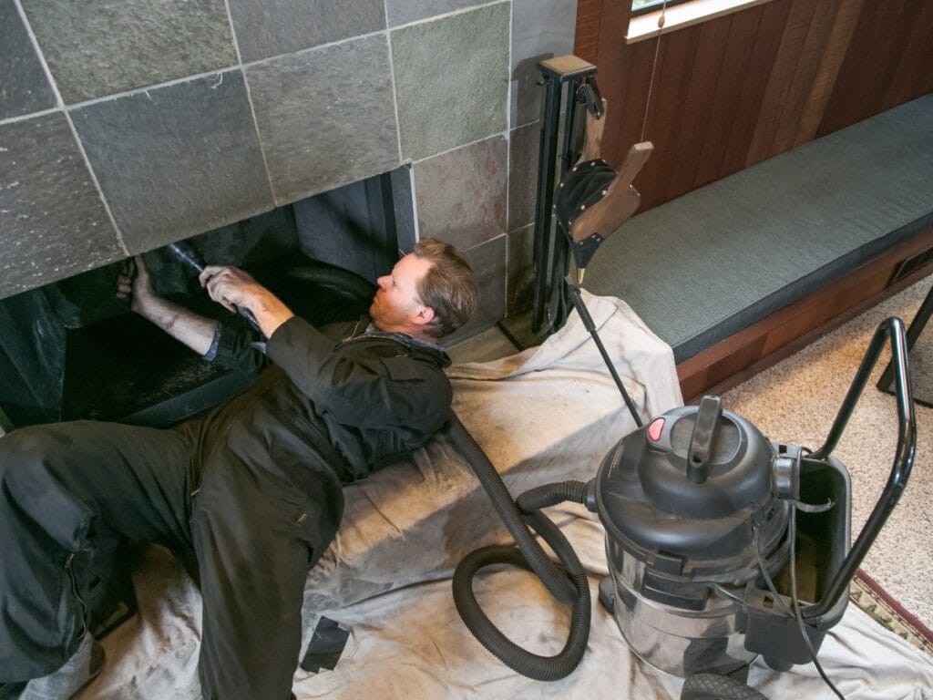 A technician in black coveralls cleans a fireplace with a vacuum while lying on a protective sheet, showcasing the meticulous care often involved in chimney maintenance and repairs like those described in Tuckpointing Explained.