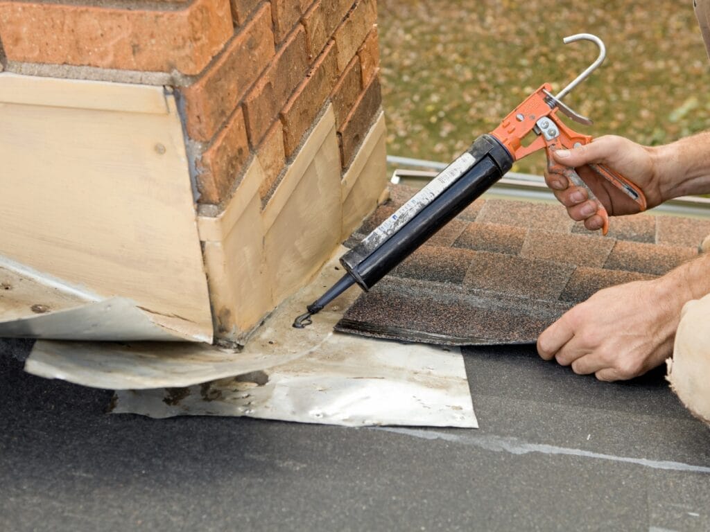 A person applies sealant with a caulking gun to metal flashing around a brick chimney on a shingled roof—an important step in chimney care, as seen in Tuckpointing Explained.